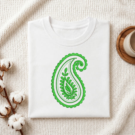 A folded white T-shirt with a green paisley leaf design on the front, styled on a textured cream fabric with cotton bolls and a small cup in the background.