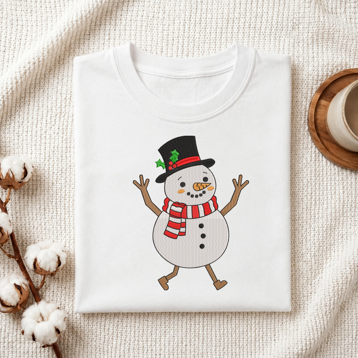 A folded white crewneck t-shirt featuring a cartoon snowman graphic with a black top hat, red and white striped scarf, and twig arms, styled on a textured cream fabric with cotton bolls and a wooden cup.