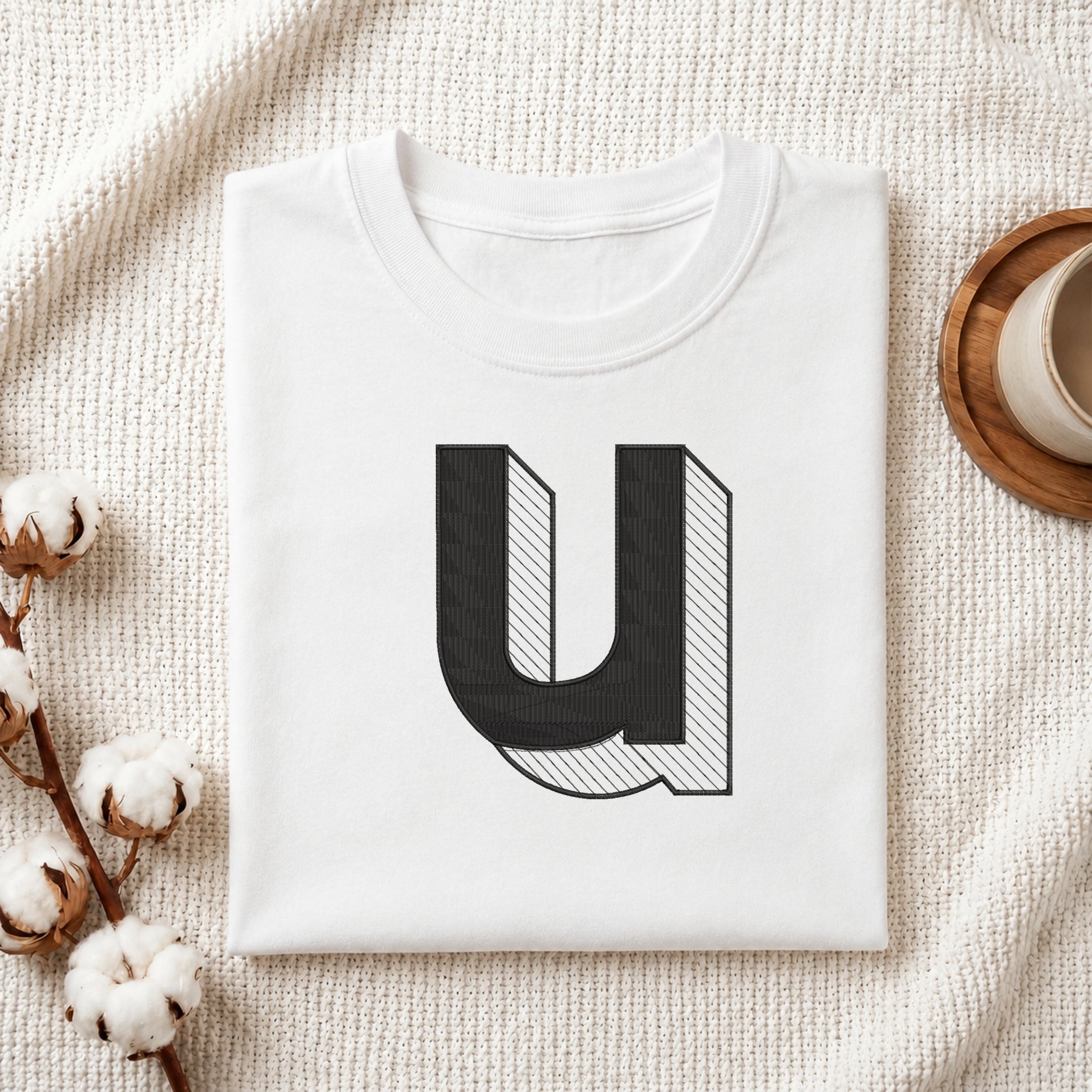 A folded white t-shirt with a large black and white graphic letter 'U' printed on the front, styled on a textured cream knit blanket with cotton stems and a cup of coffee in the background.
