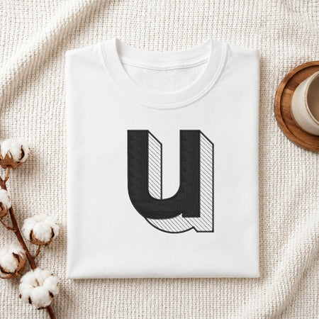 A folded white t-shirt with a large black and white graphic letter 'U' printed on the front, styled on a textured cream knit blanket with cotton stems and a cup of coffee in the background.