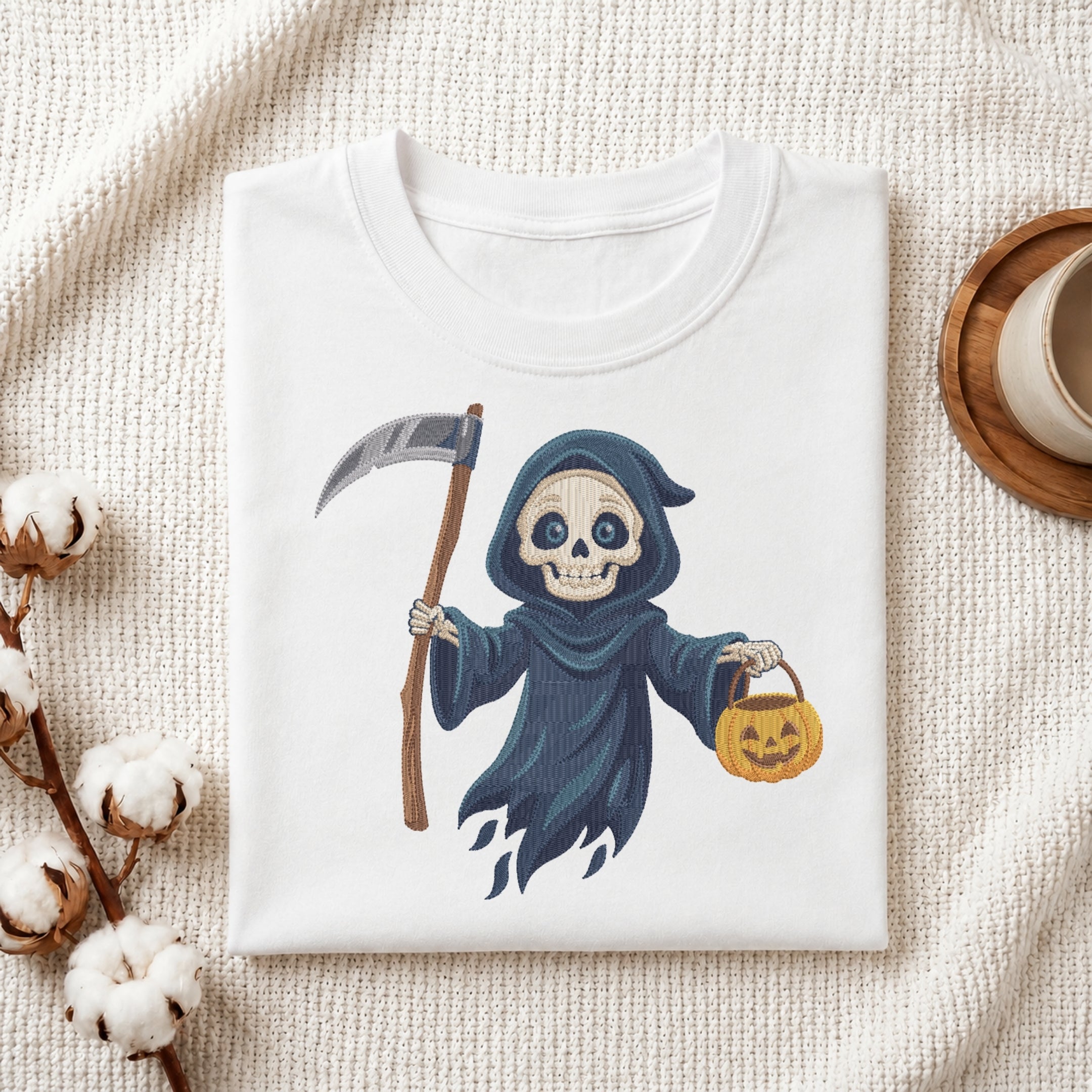 A folded white T-shirt with a cartoon grim reaper graphic, styled on a textured cream fabric with cotton bolls and a small cup in the background.