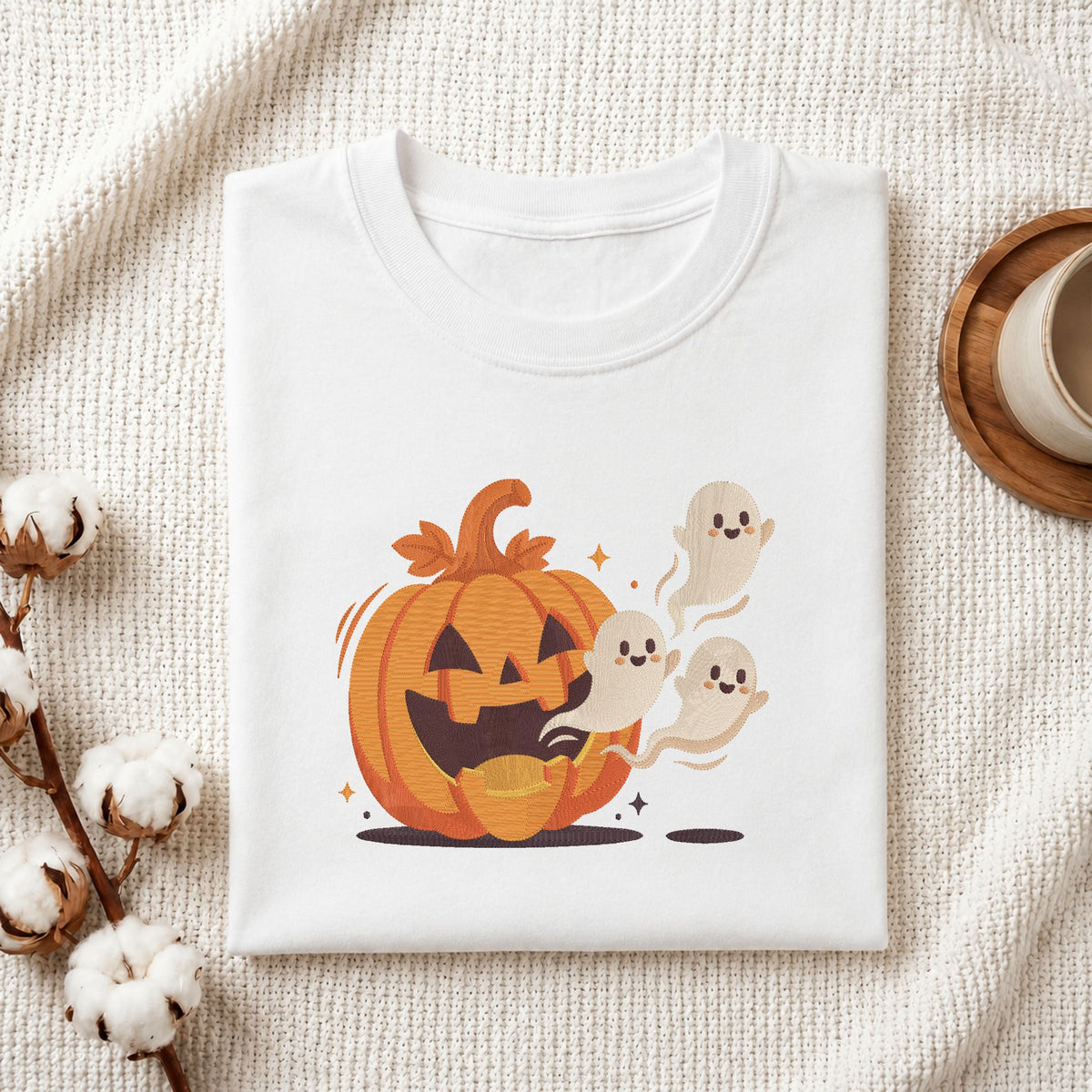 Folded white cotton t-shirt mockup featuring a cute Halloween pumpkin and ghosts machine embroidery design, styled with cotton stems and a wooden cup.