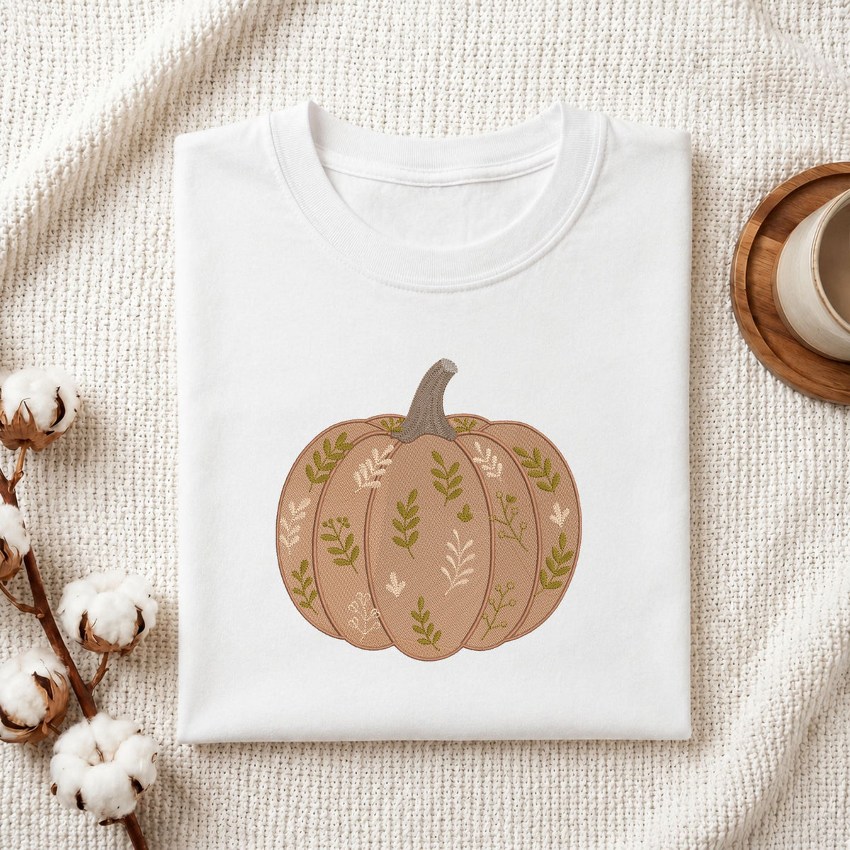  Folded white cotton t-shirt mockup featuring a leaf-patterned pumpkin machine embroidery design, styled with cotton stems and a wooden cup.   