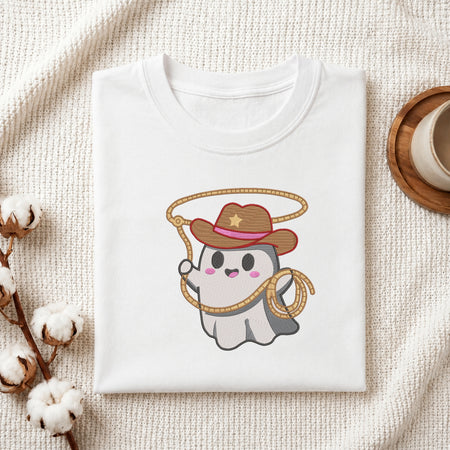 A folded white t-shirt with a cute cartoon ghost cowboy graphic, styled on a textured cream knit blanket with cotton stems and a coffee cup in the background.