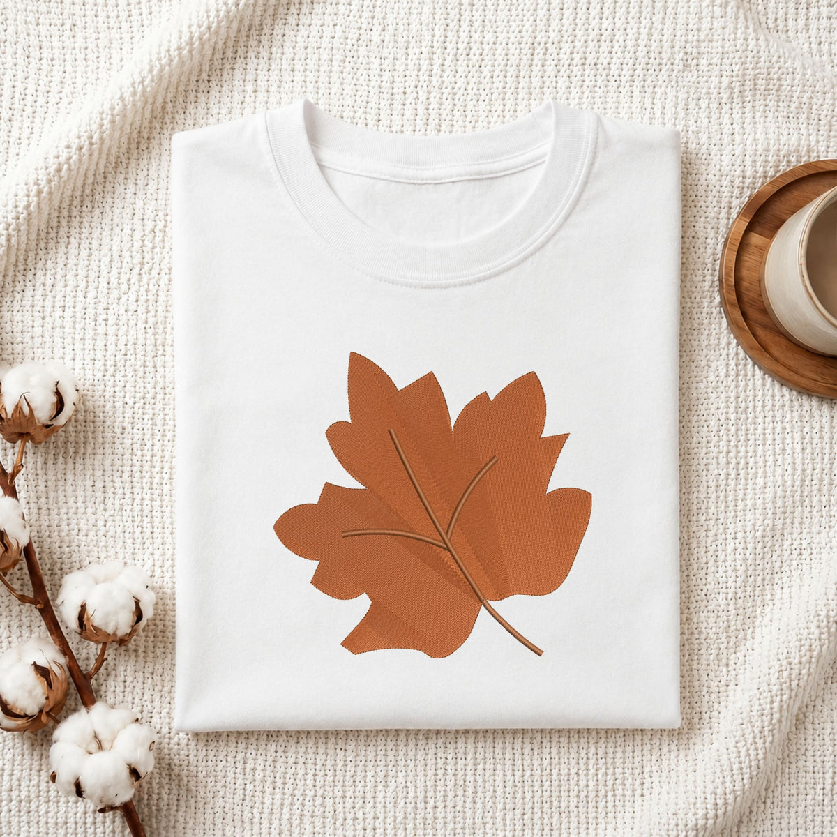 Folded white cotton t-shirt mockup featuring a brown autumn leaf machine embroidery design, styled with cotton stems and a wooden cup.