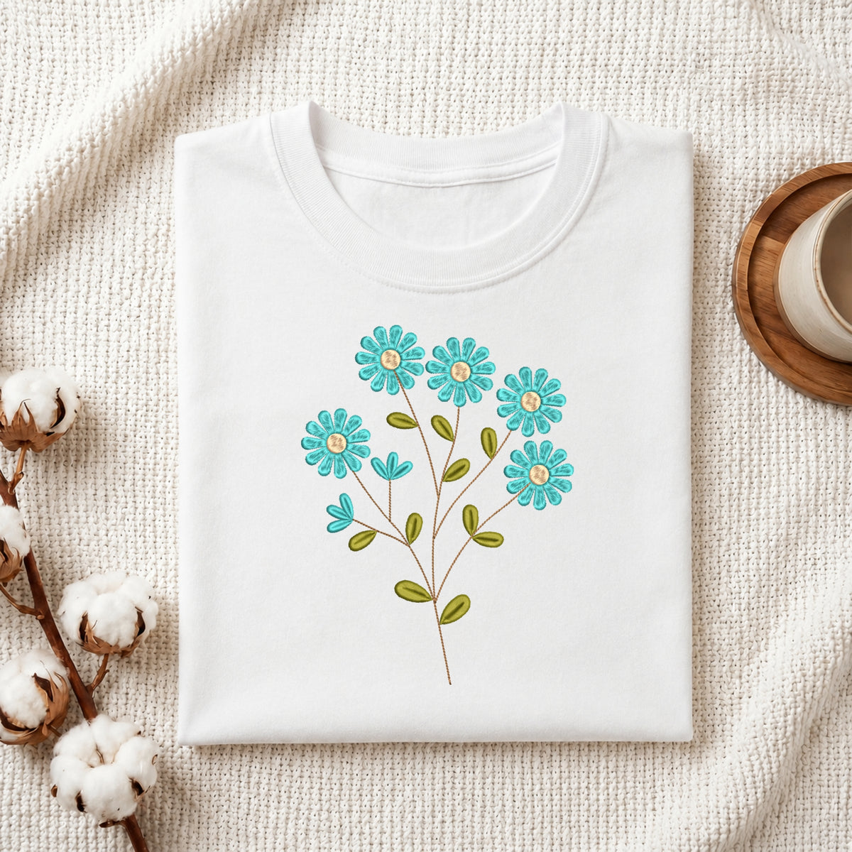 A folded white crewneck t-shirt with a colorful embroidered floral design of blue flowers and green leaves, styled on a textured cream knit blanket with cotton bolls and a wooden cup in the background.