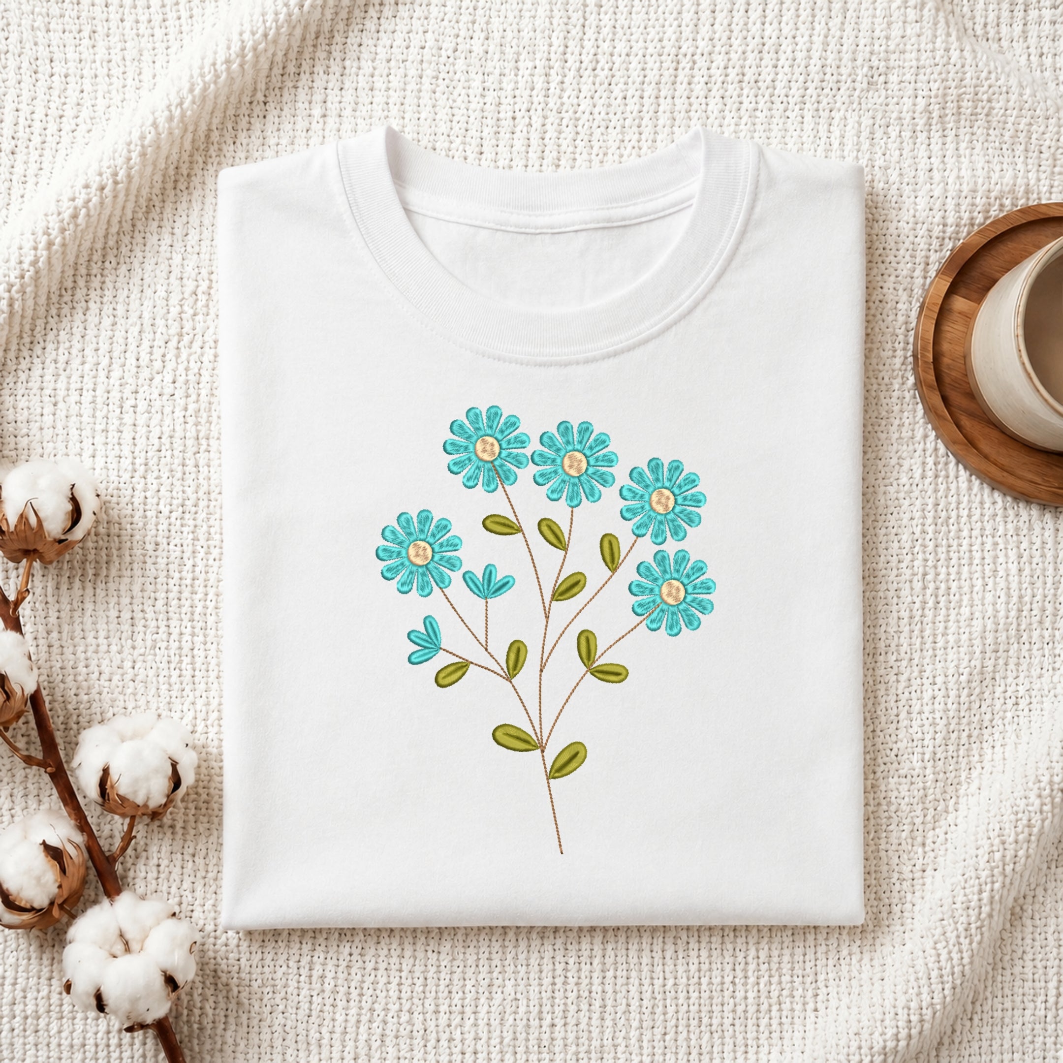 A folded white crewneck t-shirt with a colorful embroidered floral design of blue flowers and green leaves, styled on a textured cream knit blanket with cotton bolls and a wooden cup in the background.