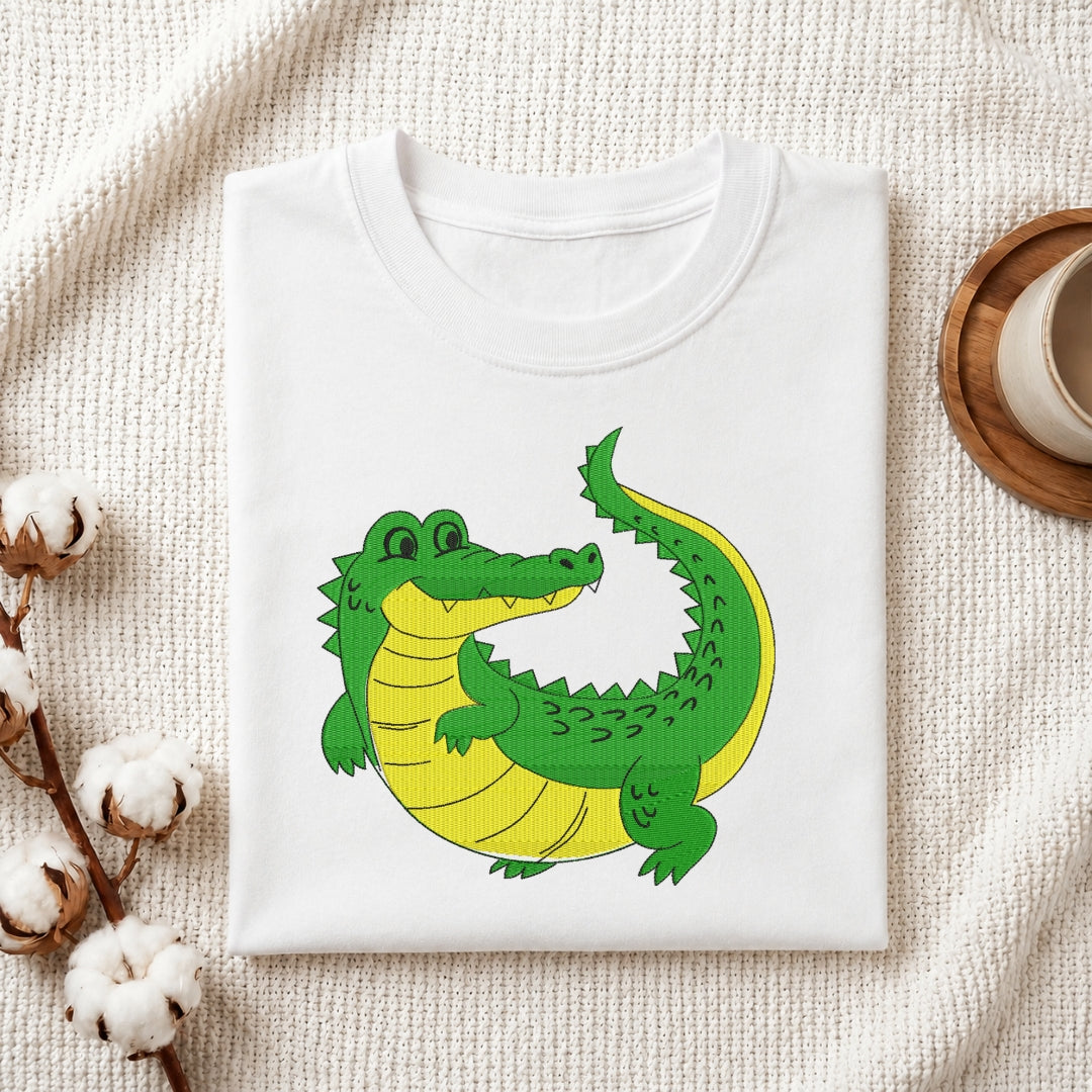 A folded white crewneck t-shirt featuring a cartoon illustration of a green and yellow crocodile, styled on a textured cream fabric with cotton bolls and a wooden cup.