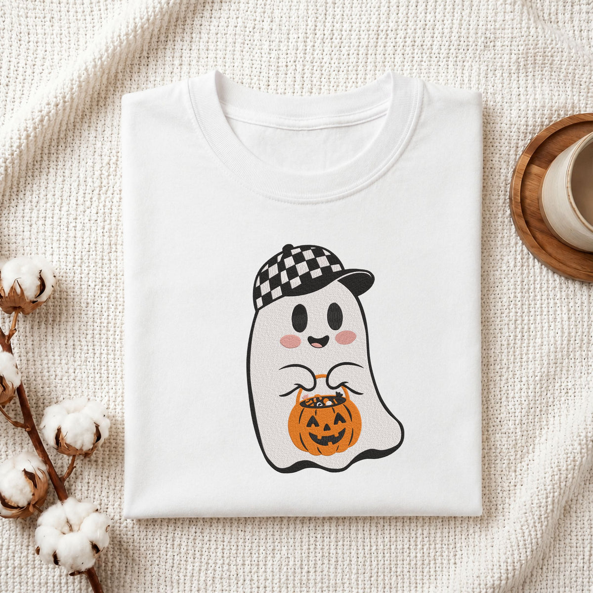 Folded white cotton t-shirt mockup featuring a cute ghost wearing a checkered baseball cap holding a pumpkin treat bucket machine embroidery design, styled with cotton stems and a wooden cup