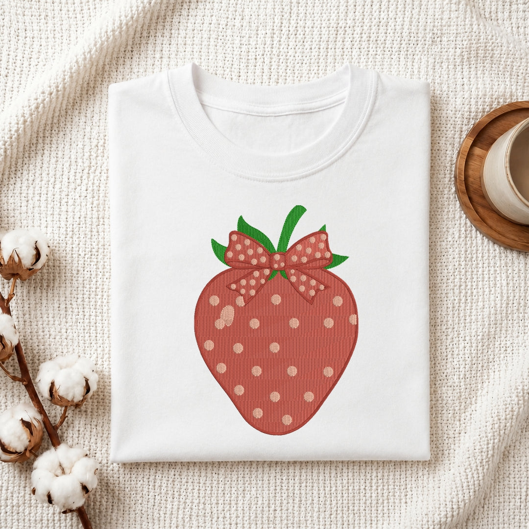 A folded white crewneck sweatshirt with a graphic of a strawberry wearing a polka-dot bow, styled on a textured cream fabric with cotton bolls and a wooden cup.