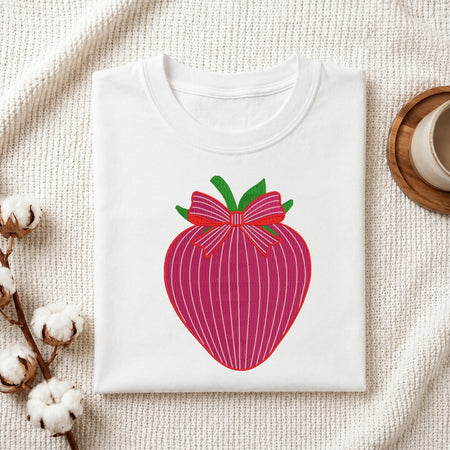 A folded white crewneck t-shirt featuring a stylized graphic of a pink striped strawberry with green leaves and a red bow, displayed on a textured cream fabric background with cotton bolls and a wooden cup.