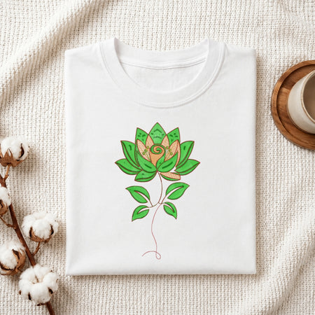 A folded white crewneck t-shirt with a green and gold lotus flower graphic on the front, styled on a textured cream knit blanket with cotton bolls and a wooden cup in the background.