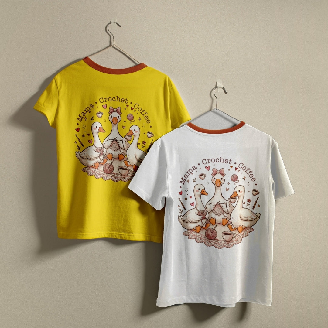 Two t-shirts hanging on a wall, one bright yellow and one white, both featuring a round colorful illustration of cozy cartoon ducks crocheting.