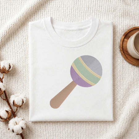 A folded white t-shirt with a pastel-colored pacifier/rattle graphic, styled on a textured cream knit blanket with cotton bolls and a wooden cup.