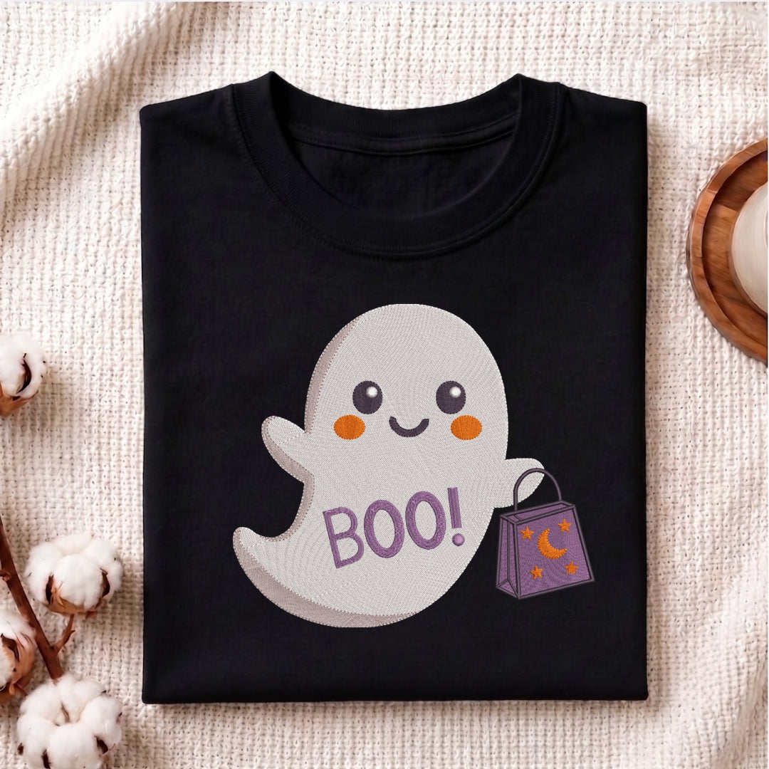 A folded black t-shirt with a cute cartoon ghost graphic holding a purple trick-or-treat bag with 'BOO!' written on it, displayed on a textured beige fabric background with cotton bolls.
