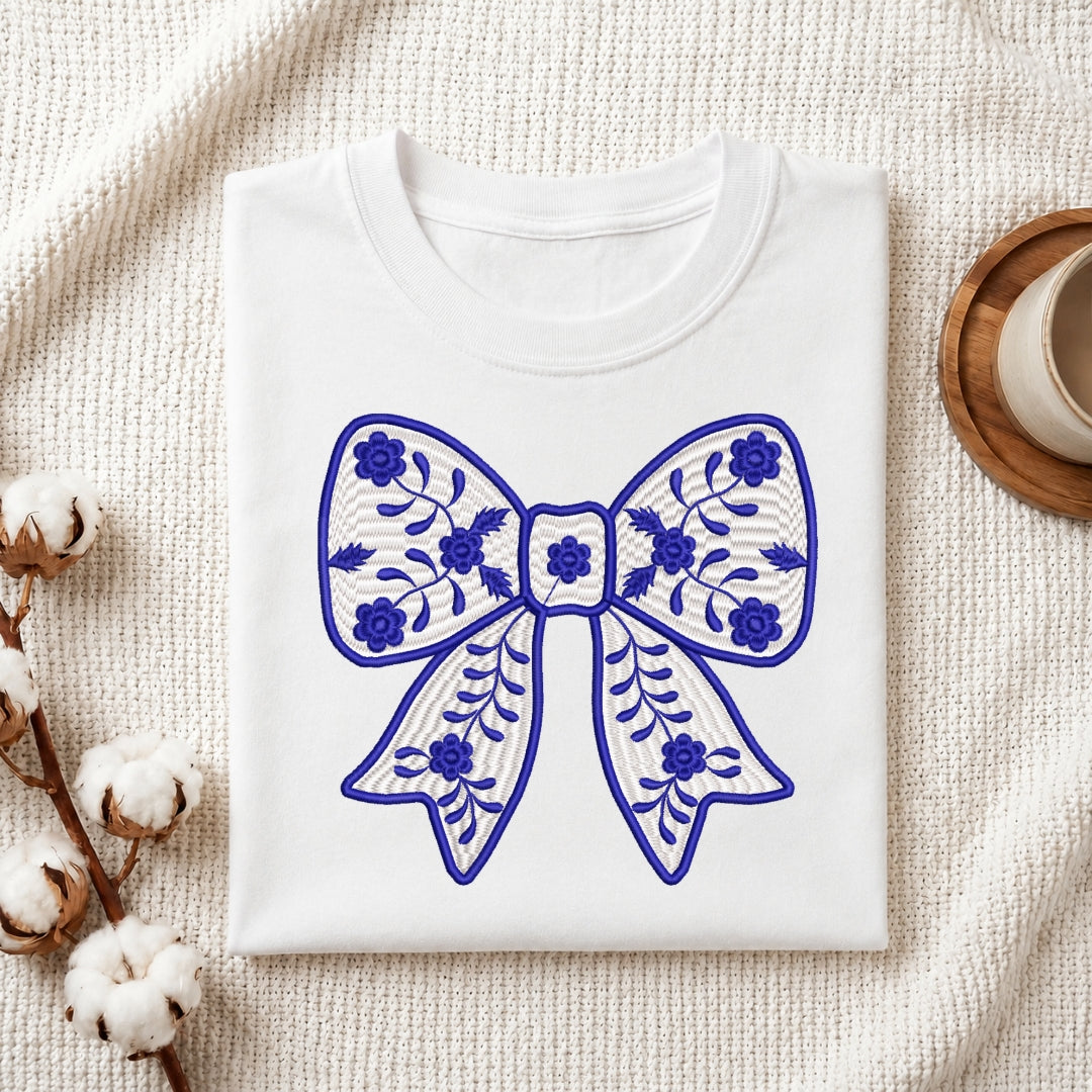 A folded white crewneck sweatshirt or t-shirt with a blue embroidered bow design on the front, styled on a textured cream fabric background with cotton bolls and a wooden cup.