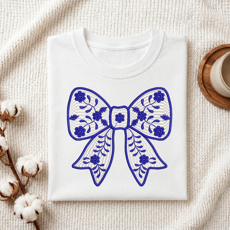 A folded white crewneck sweatshirt or t-shirt with a blue embroidered bow design on the front, styled on a textured cream fabric background with cotton bolls and a wooden cup.