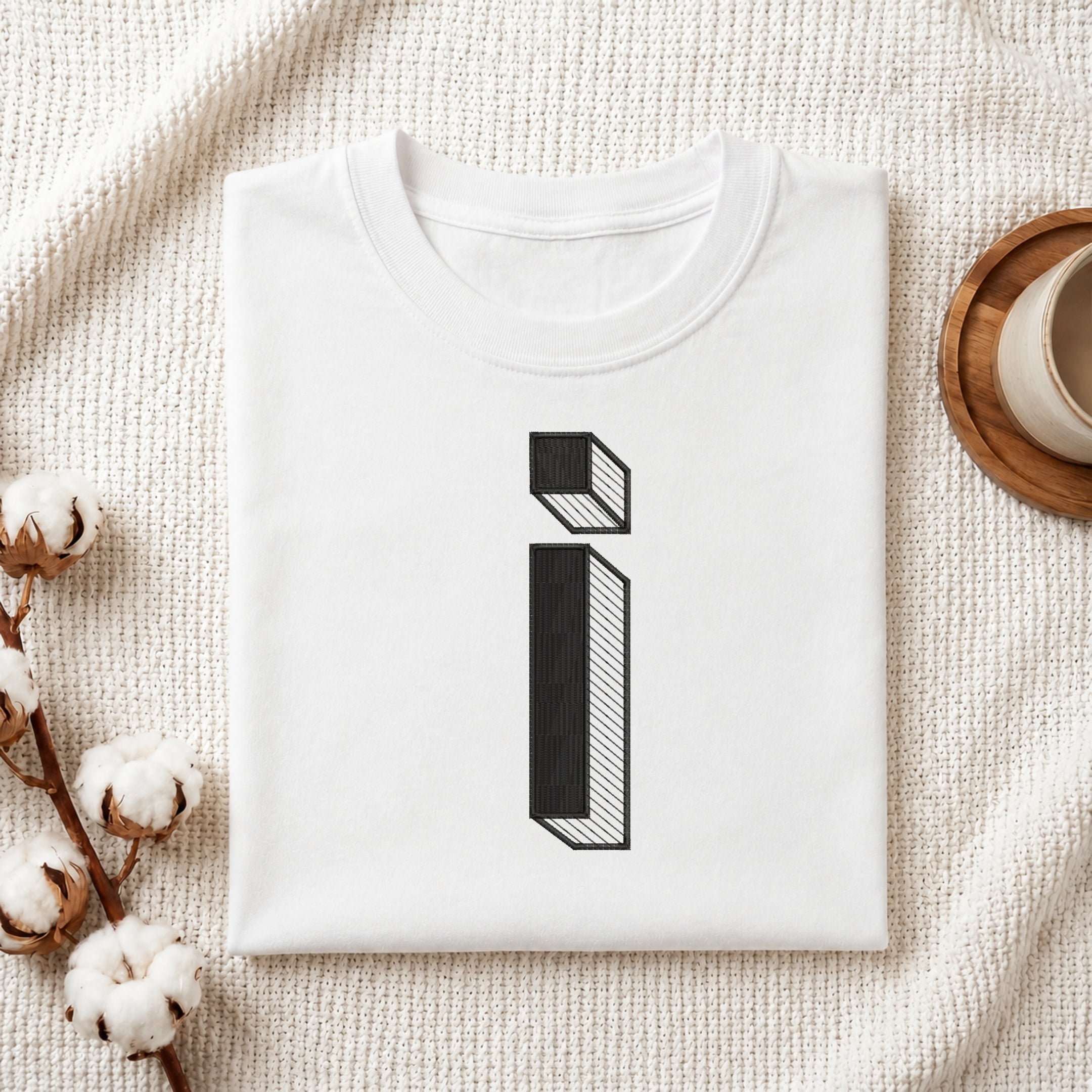 A folded white t-shirt with a black 3D-effect letter 'i' graphic, styled on a textured cream fabric with cotton bolls and a wooden cup in the background.