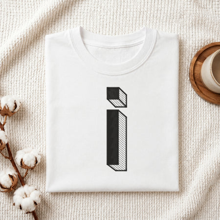 A folded white t-shirt with a black 3D-effect letter 'i' graphic, styled on a textured cream fabric with cotton bolls and a wooden cup in the background.