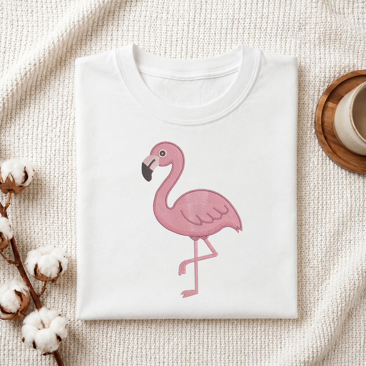 A folded white crewneck t-shirt featuring a pink flamingo graphic on the front, styled on a textured cream fabric with cotton bolls and a wooden cup in the background.