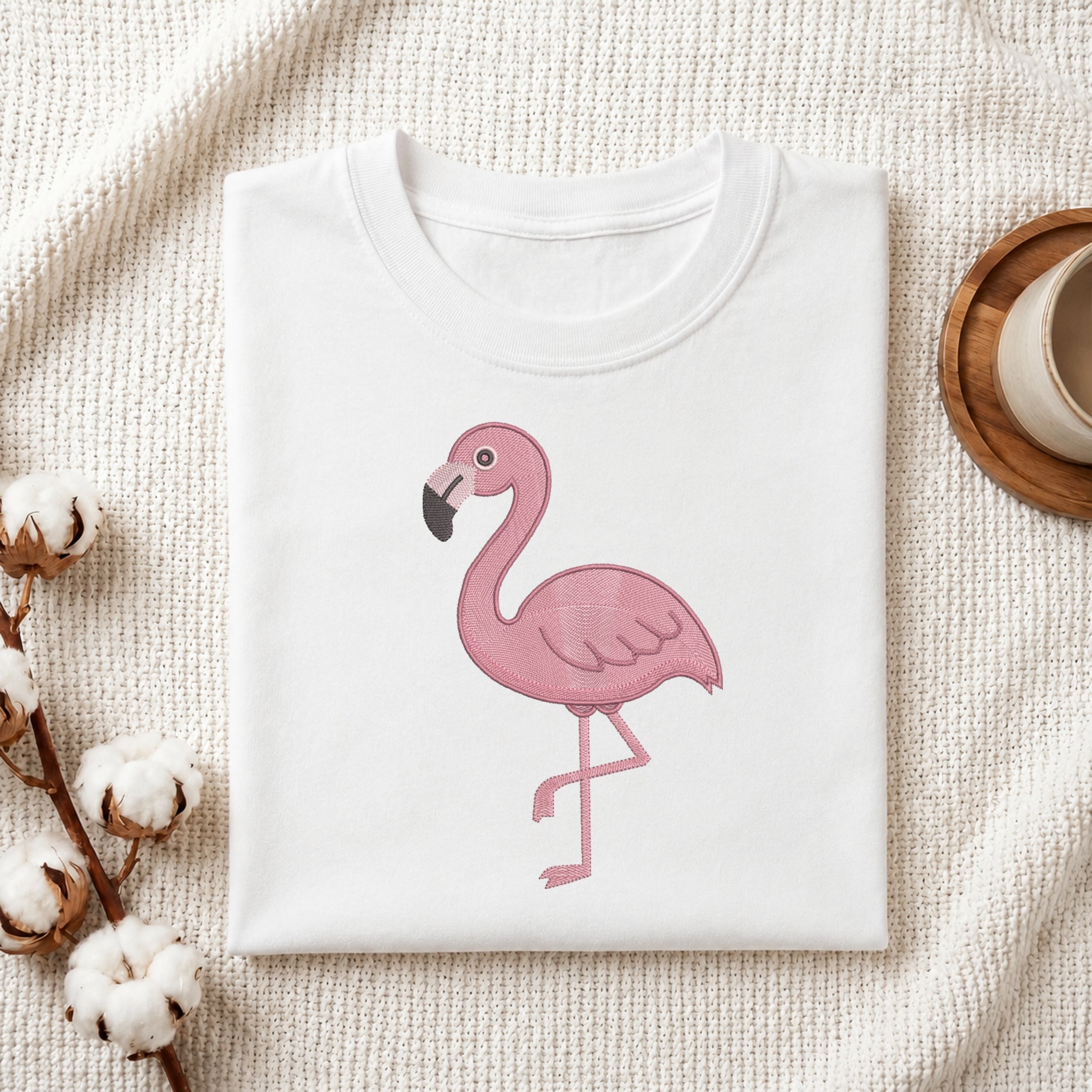 A folded white crewneck t-shirt featuring a pink flamingo graphic on the front, styled on a textured cream fabric with cotton bolls and a wooden cup in the background.