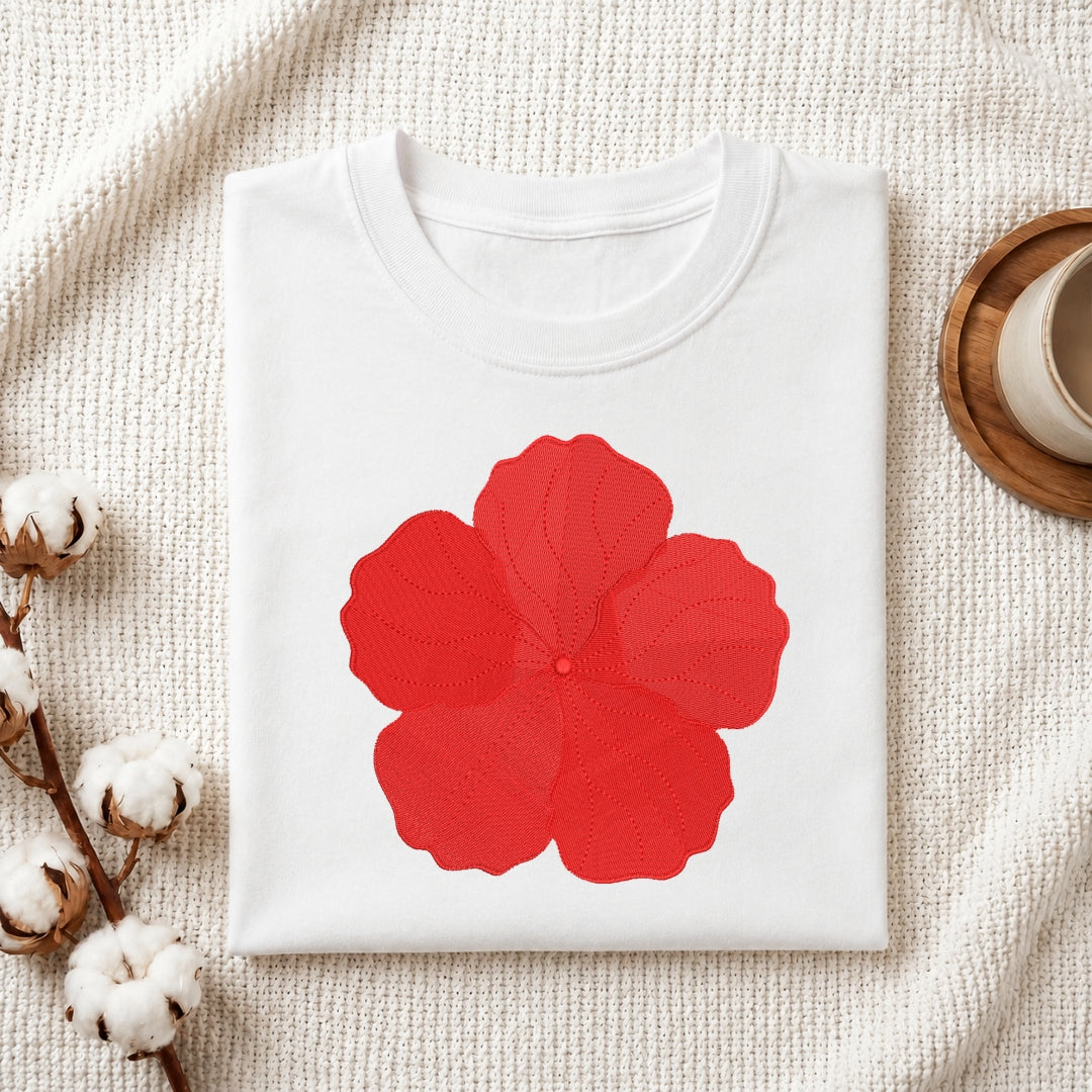 A folded white crewneck t-shirt with a large red flower graphic on the front, styled on a textured cream fabric background with cotton bolls and a wooden cup.