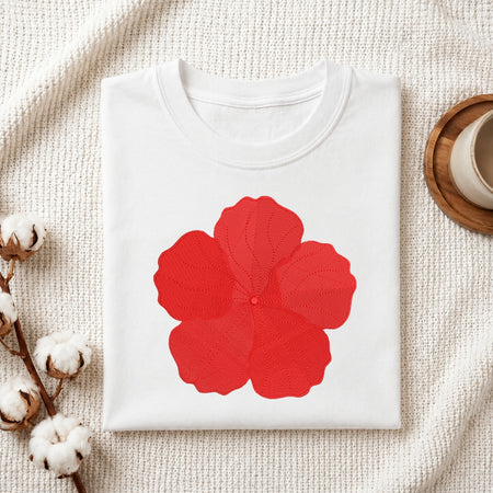 A folded white crewneck t-shirt with a large red flower graphic on the front, styled on a textured cream fabric background with cotton bolls and a wooden cup.