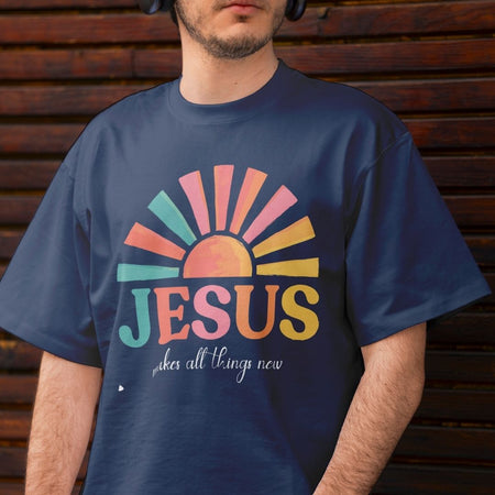 A man wearing a navy blue t-shirt with a colorful retro-style sun graphic and the text 'JESUS makes all things new' printed on the front, set against a dark wooden plank background.