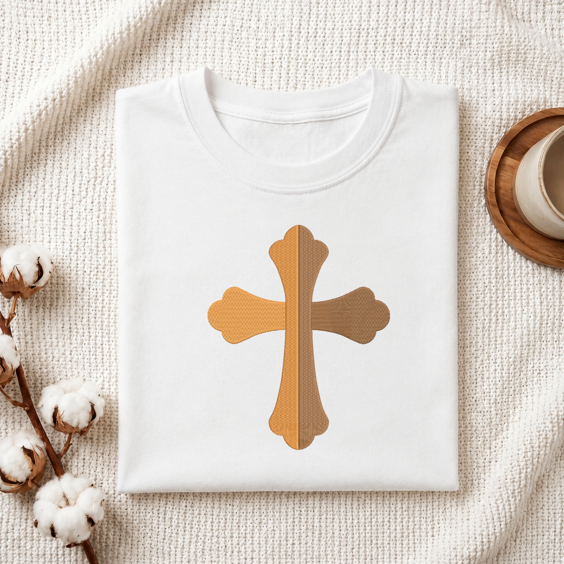 A folded white T-shirt laid flat on a textured cream fabric background, featuring a central cross graphic in warm tan and brown woodgrain textures. Cotton bolls and part of a wooden cup are visible as styled props.