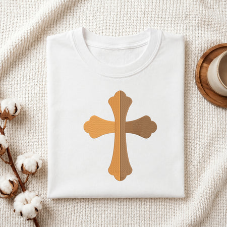 A folded white T-shirt laid flat on a textured cream fabric background, featuring a central cross graphic in warm tan and brown woodgrain textures. Cotton bolls and part of a wooden cup are visible as styled props.