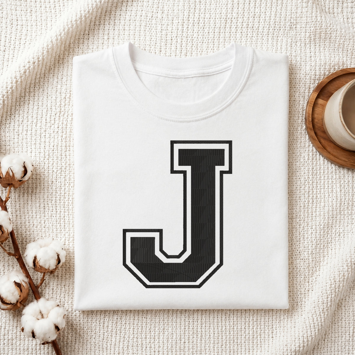 A folded white T-shirt with a large black block letter J outlined in white on the front, styled on a textured cream fabric with cotton bolls and a cup in the background.