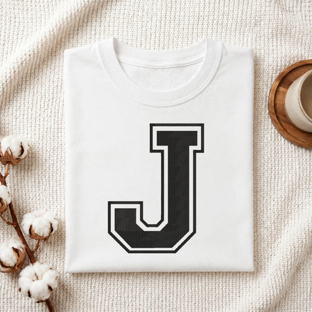 A folded white T-shirt with a large black block letter J outlined in white on the front, styled on a textured cream fabric with cotton bolls and a cup in the background.