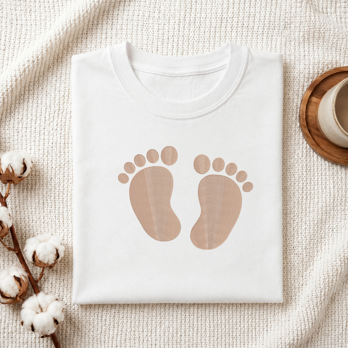 A folded white T-shirt with a pair of tan baby footprints printed on the front, styled on a textured cream blanket with cotton stems and a cup in the background.