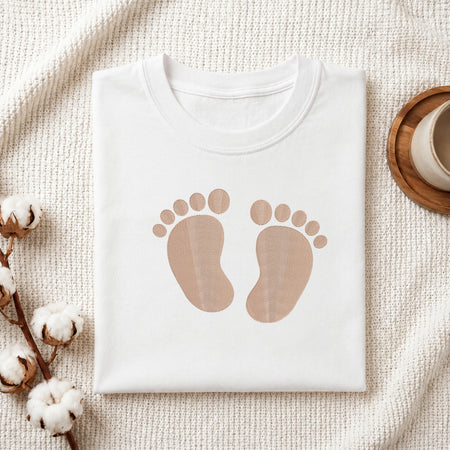 A folded white T-shirt with a pair of tan baby footprints printed on the front, styled on a textured cream blanket with cotton stems and a cup in the background.
