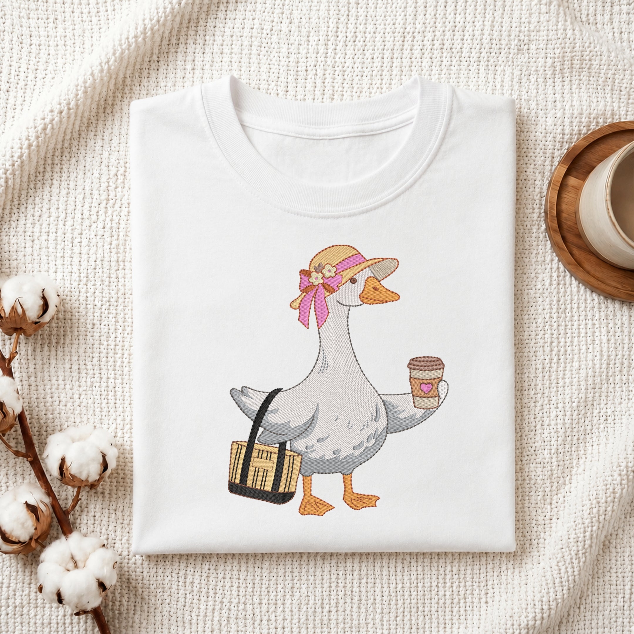 A folded white crewneck t-shirt featuring a cartoon graphic of a goose wearing a straw hat with a pink bow, carrying a black and yellow bag and holding a coffee cup. The t-shirt is styled in a flat-lay on a textured cream fabric with cotton bolls and a wooden cup on a coaster.