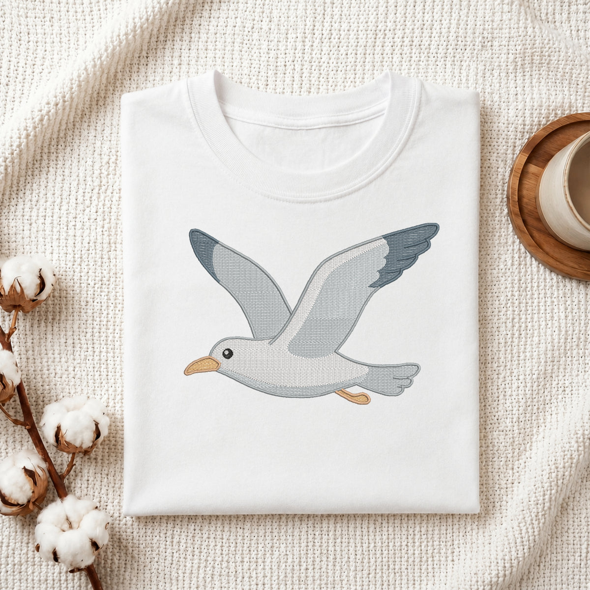 A folded white crewneck t-shirt featuring an embroidered flying seagull design, styled on a textured cream knit blanket with cotton bolls and a wooden cup in the background.