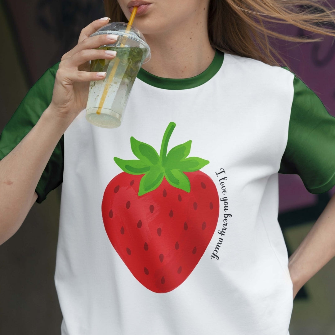 Person wearing a white t-shirt with a strawberry graphic and green sleeves, holding a drink.