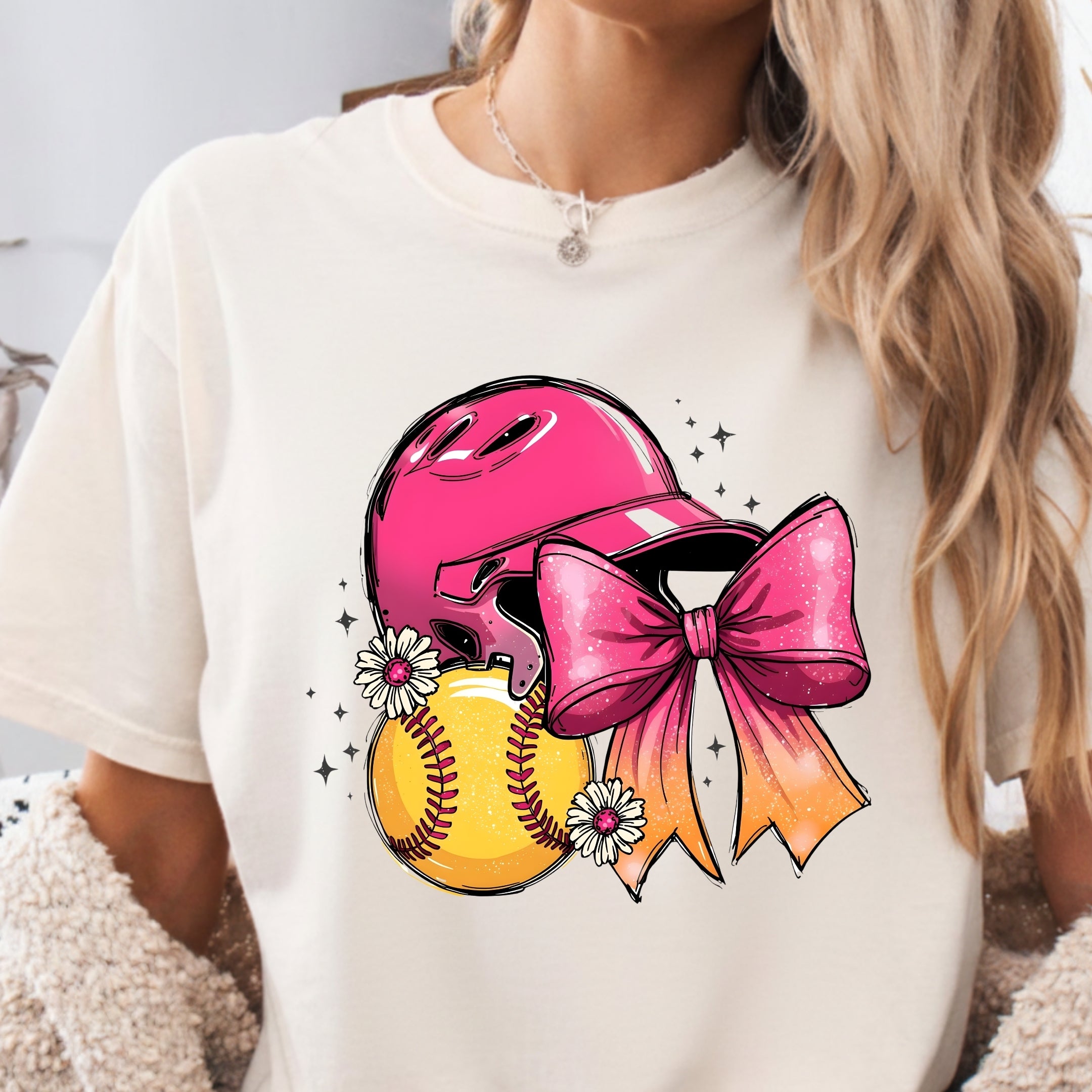 A woman wearing a cream-colored t-shirt with a colorful graphic of a pink baseball helmet, a yellow softball, a pink and orange bow, and small white flowers.
