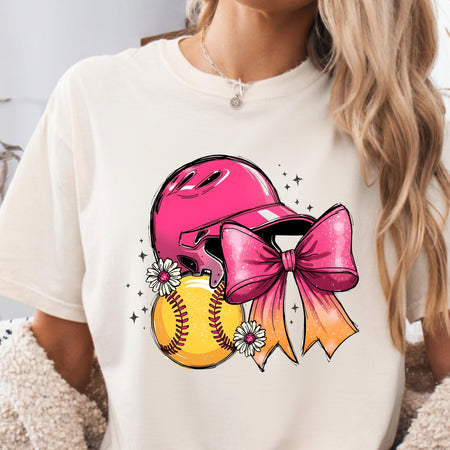 A woman wearing a cream-colored t-shirt with a colorful graphic of a pink baseball helmet, a yellow softball, a pink and orange bow, and small white flowers.