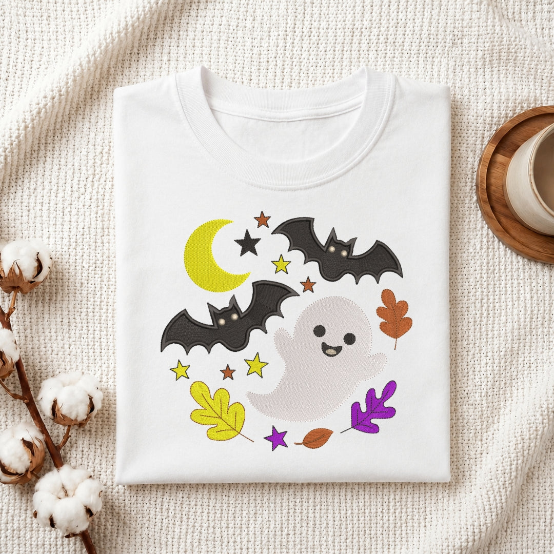 A folded white crewneck t-shirt with a cute Halloween graphic print, displayed on a textured beige fabric background with decorative cotton bolls and a wooden cup.
