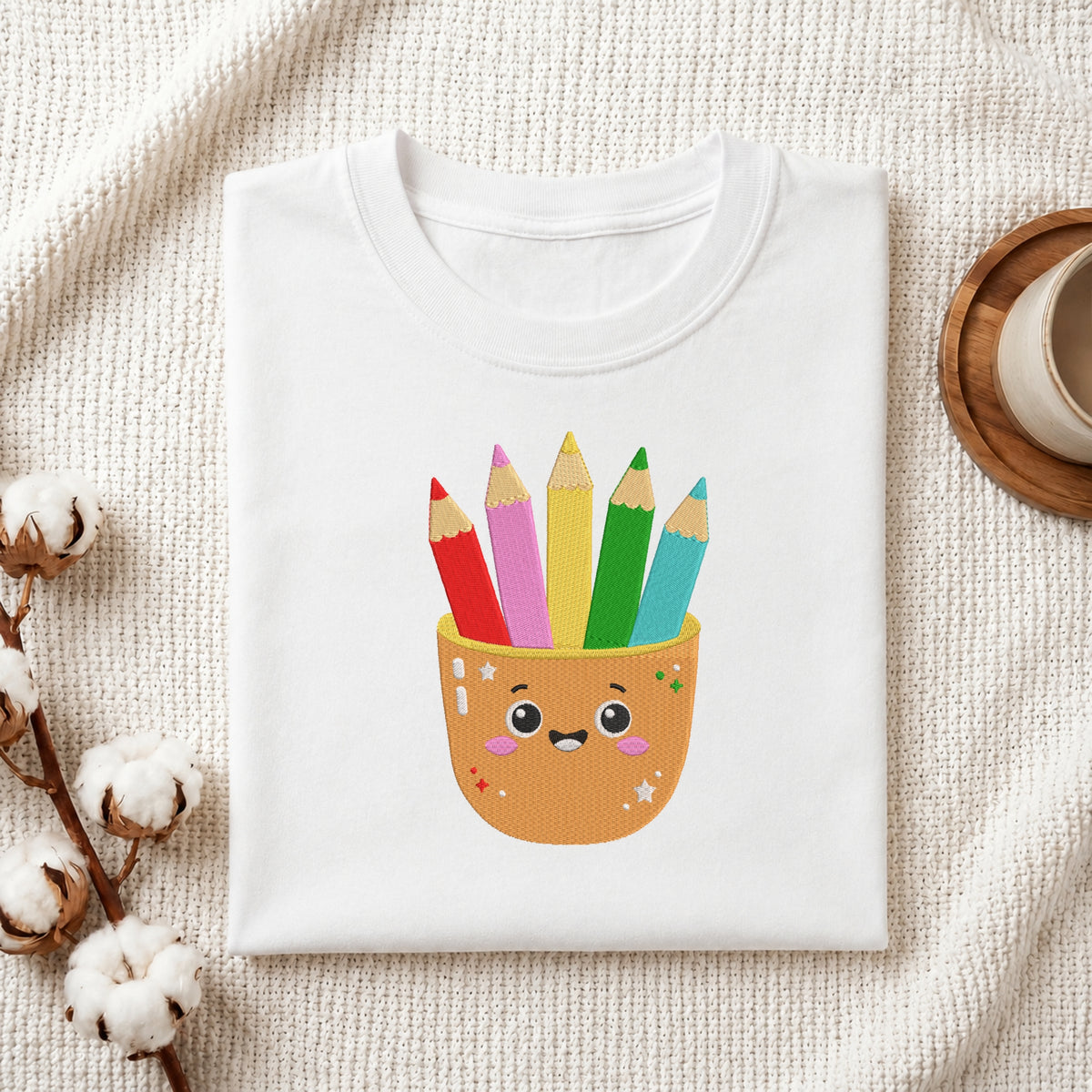 A folded white T-shirt with a cute graphic of a smiling orange pencil cup holding five colorful pencils, styled on a textured cream fabric with cotton bolls and a wooden cup in the background.