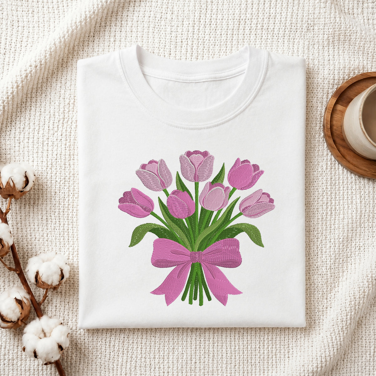 A folded white crewneck T-shirt with a graphic of pink tulips and a pink ribbon, styled on a textured cream fabric with cotton bolls and a cup on a wooden tray.