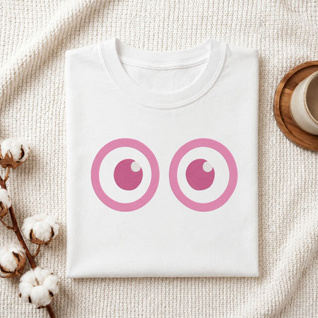 A folded white t-shirt with a pink cartoon eyes graphic printed on the front, displayed on a textured cream knit blanket with cotton bolls and a wooden cup in the background.