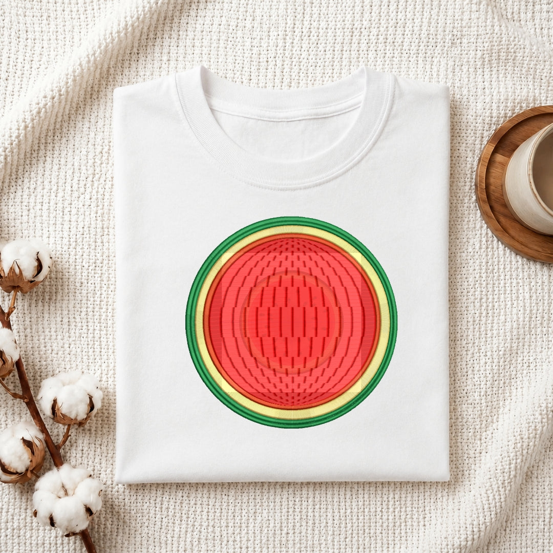 A folded white crewneck t-shirt featuring a circular watermelon graphic print, styled on a textured cream fabric background with cotton bolls and a wooden cup.