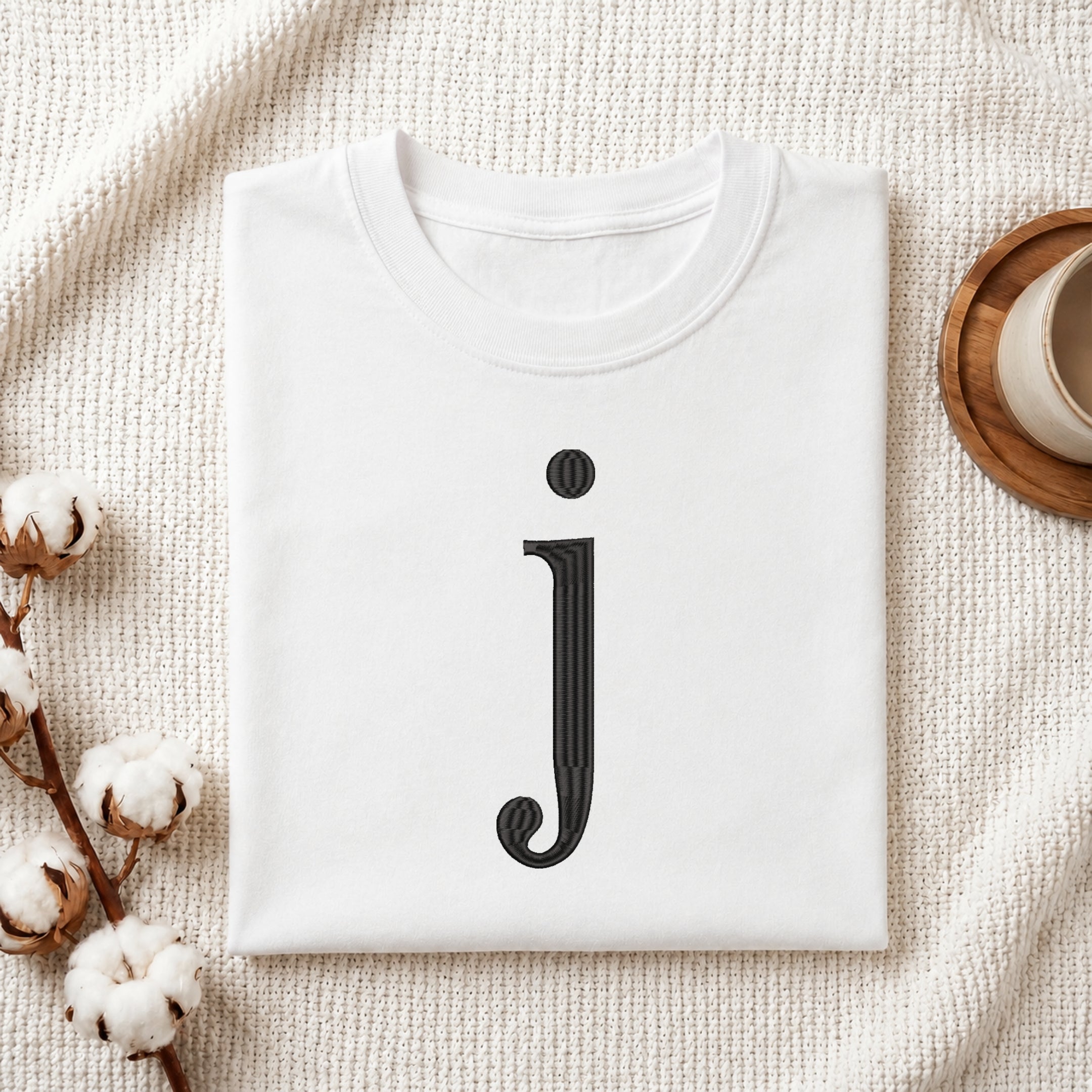 A folded white t-shirt with a black stylized lowercase letter 'j' printed on the front, displayed in a flat-lay arrangement on a textured cream fabric with cotton stems and a cup on a wooden coaster.