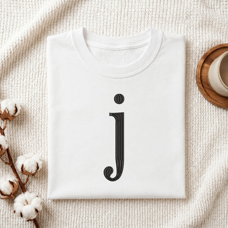 A folded white t-shirt with a black stylized lowercase letter 'j' printed on the front, displayed in a flat-lay arrangement on a textured cream fabric with cotton stems and a cup on a wooden coaster.