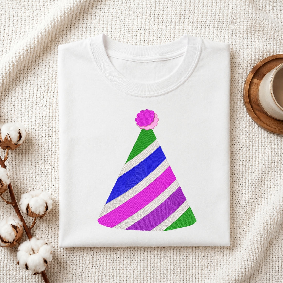 A folded white T-shirt with a colorful striped party hat graphic, displayed on a textured cream fabric background with cotton bolls and a small cup in the corners.