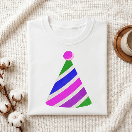 A folded white T-shirt with a colorful striped party hat graphic, displayed on a textured cream fabric background with cotton bolls and a small cup in the corners.