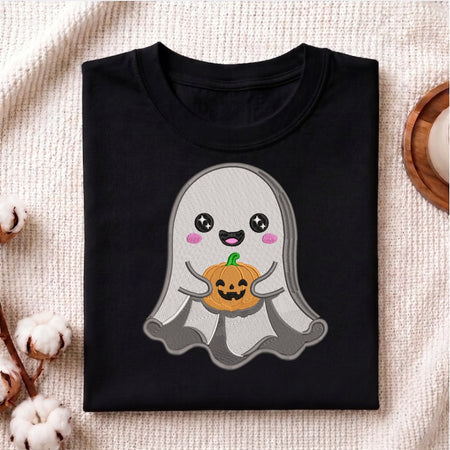 A folded black t-shirt with a cute cartoon ghost holding a jack-o'-lantern pumpkin, displayed on a textured beige fabric background with decorative cotton bolls.