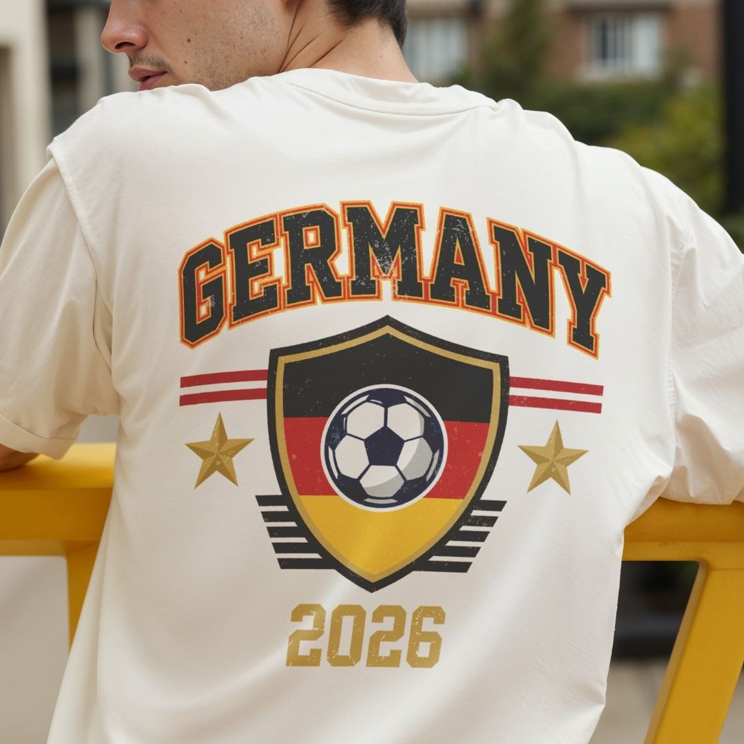 Close up of the back of a man's cream-colored t-shirt showing a large vintage style Germany soccer crest design in black, red, and gold with the text Germany 2026.